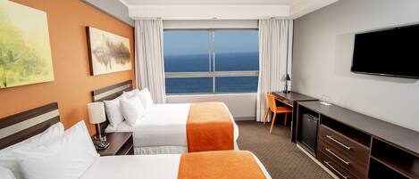 A hotel room with two beds, a desk, a TV, and a large window with a view of the sea.