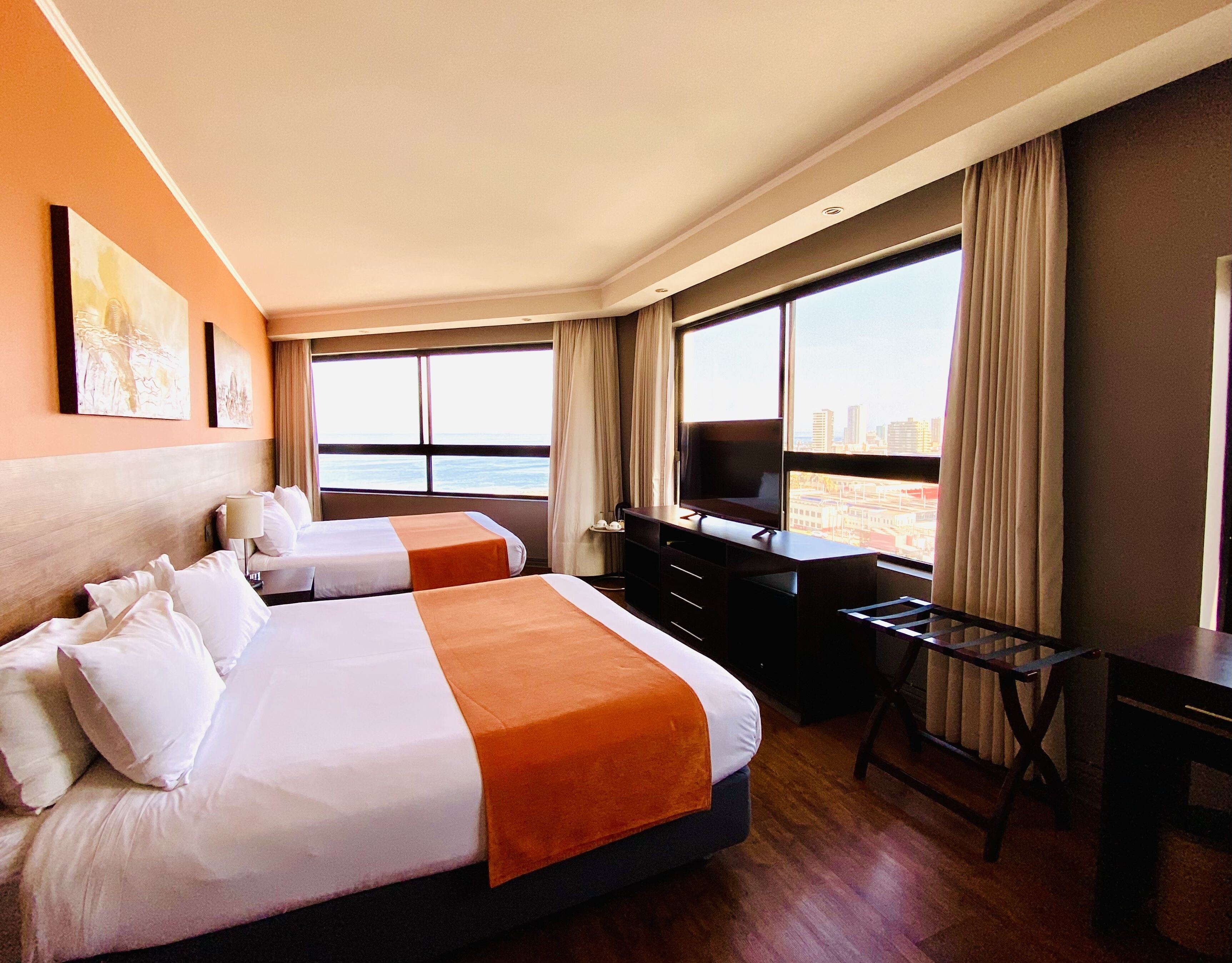 A hotel room with two beds, a desk, a TV, and large windows offering a city view.
