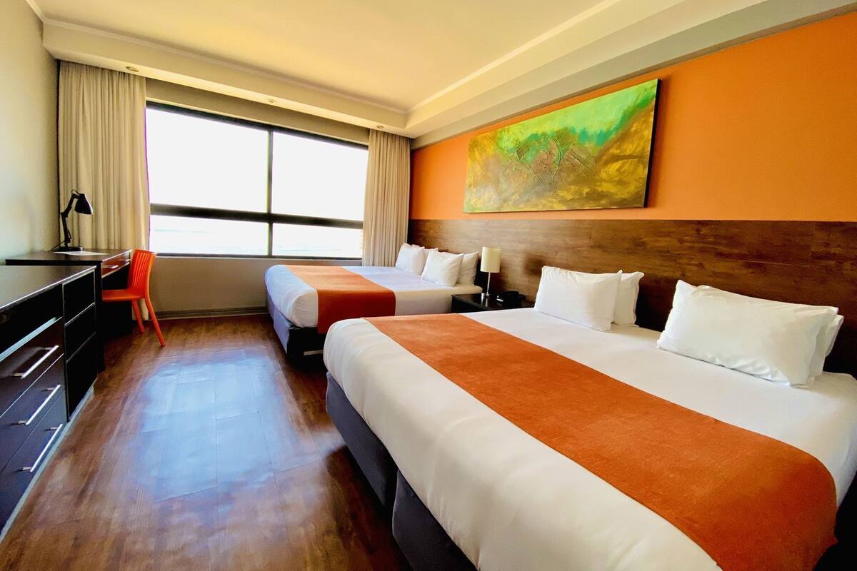 A hotel room with two beds, a desk, a chair, and a large window.