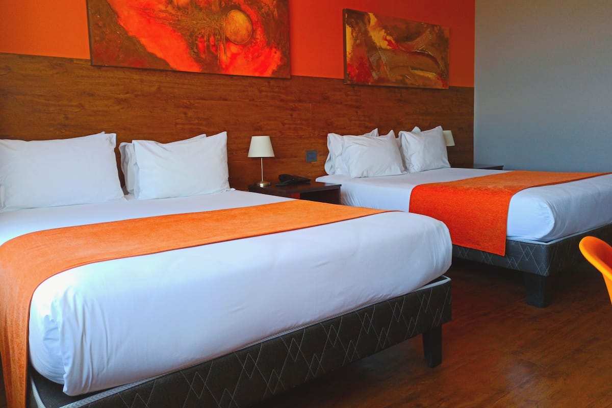 A hotel room with two beds, a wooden headboard, a nightstand, a lamp, and two paintings on the wall.