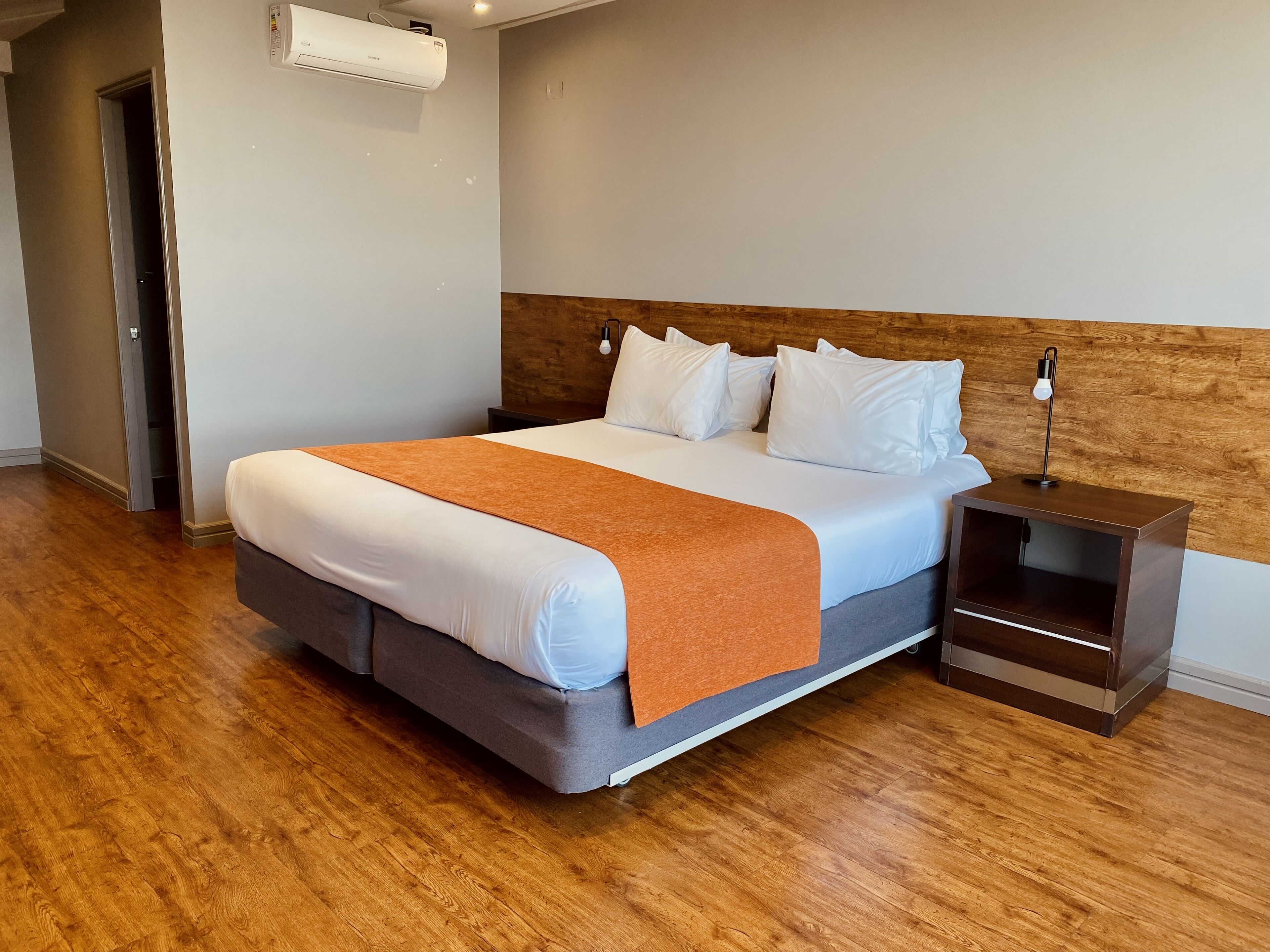 A modern hotel room with a large bed, a wooden headboard, a nightstand, and a built-in shelf.