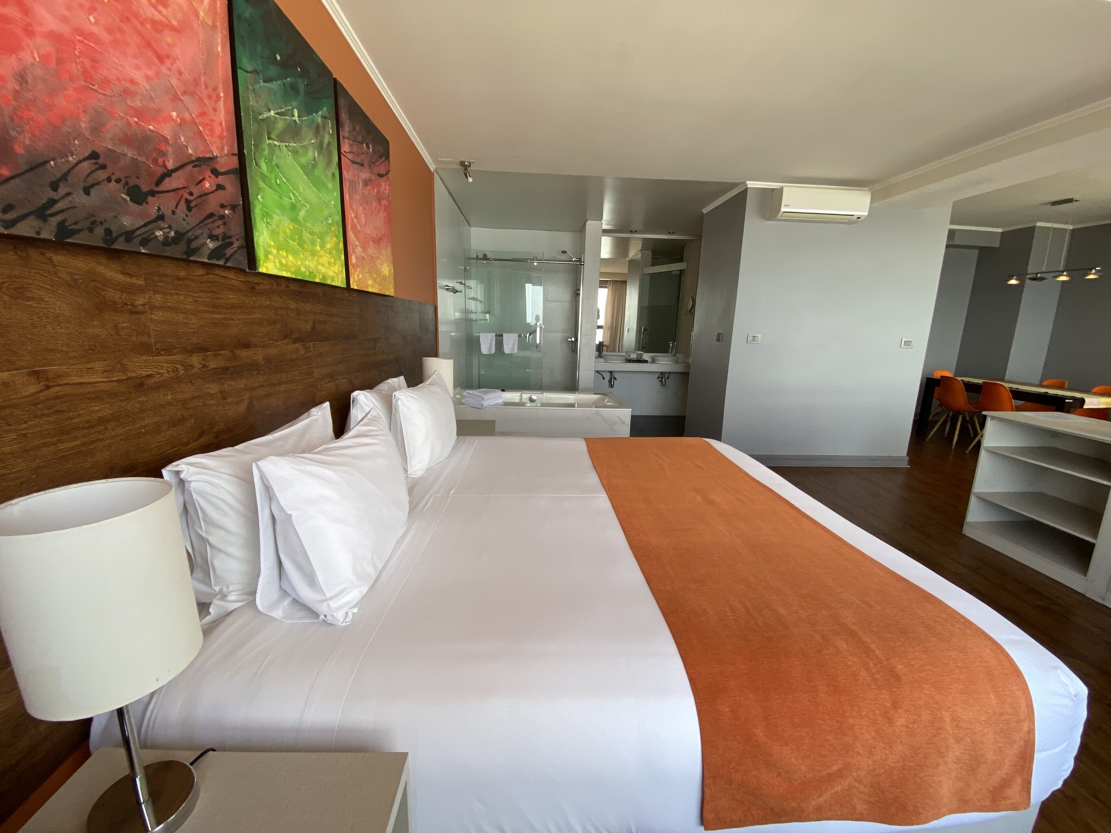 Habitación de hotel con una cama grande, una mesita de noche con lámpara, un baño con cabina de ducha de vidrio y un área de comedor con sillas naranjas.