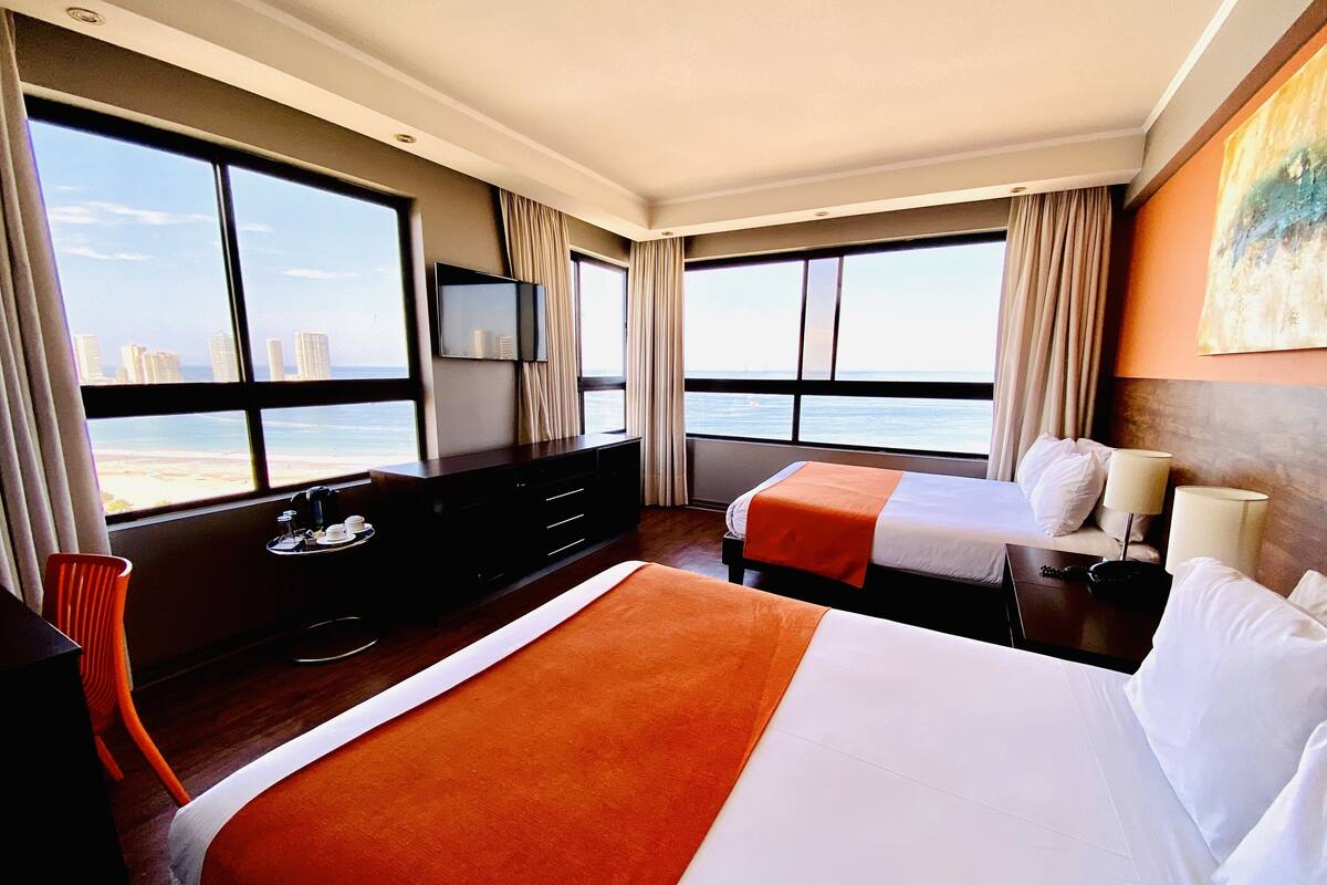 A hotel room with two beds, a TV, a desk, and a view of the beach.