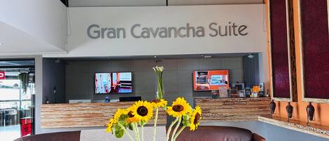 A hotel lobby with a large sign reading "Gran Cavancha Suite," a reception desk, a brown leather sofa, a vase with sunflowers, and hanging wicker lamps.