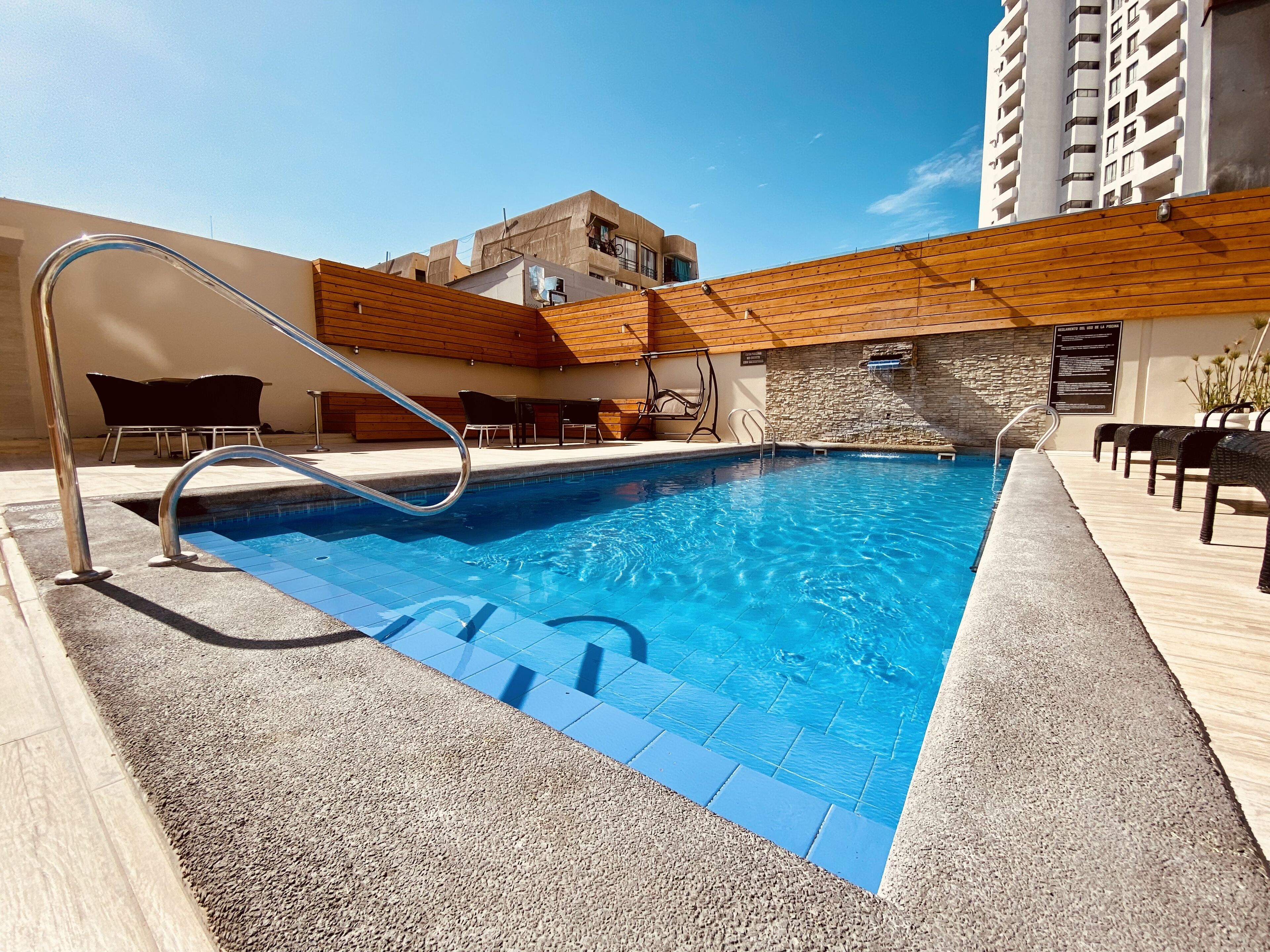 Una piscina con una escalera metálica, rodeada de sillas de descanso y un muro de piedra.