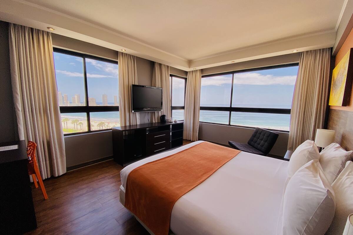 A hotel room with a large bed, a TV, a desk, and a view of the beach and city skyline.