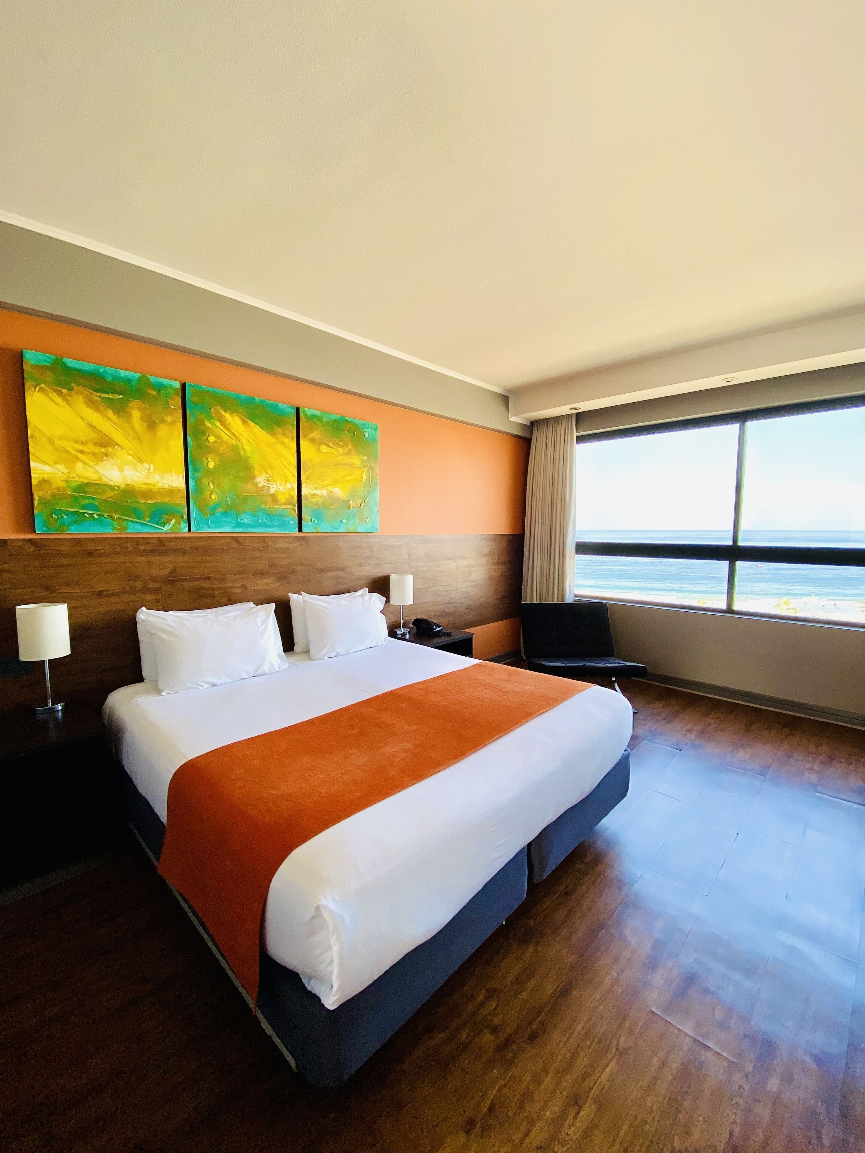 A hotel room with a large bed, a nightstand, a lamp, a chair, and a window with a view of the ocean.