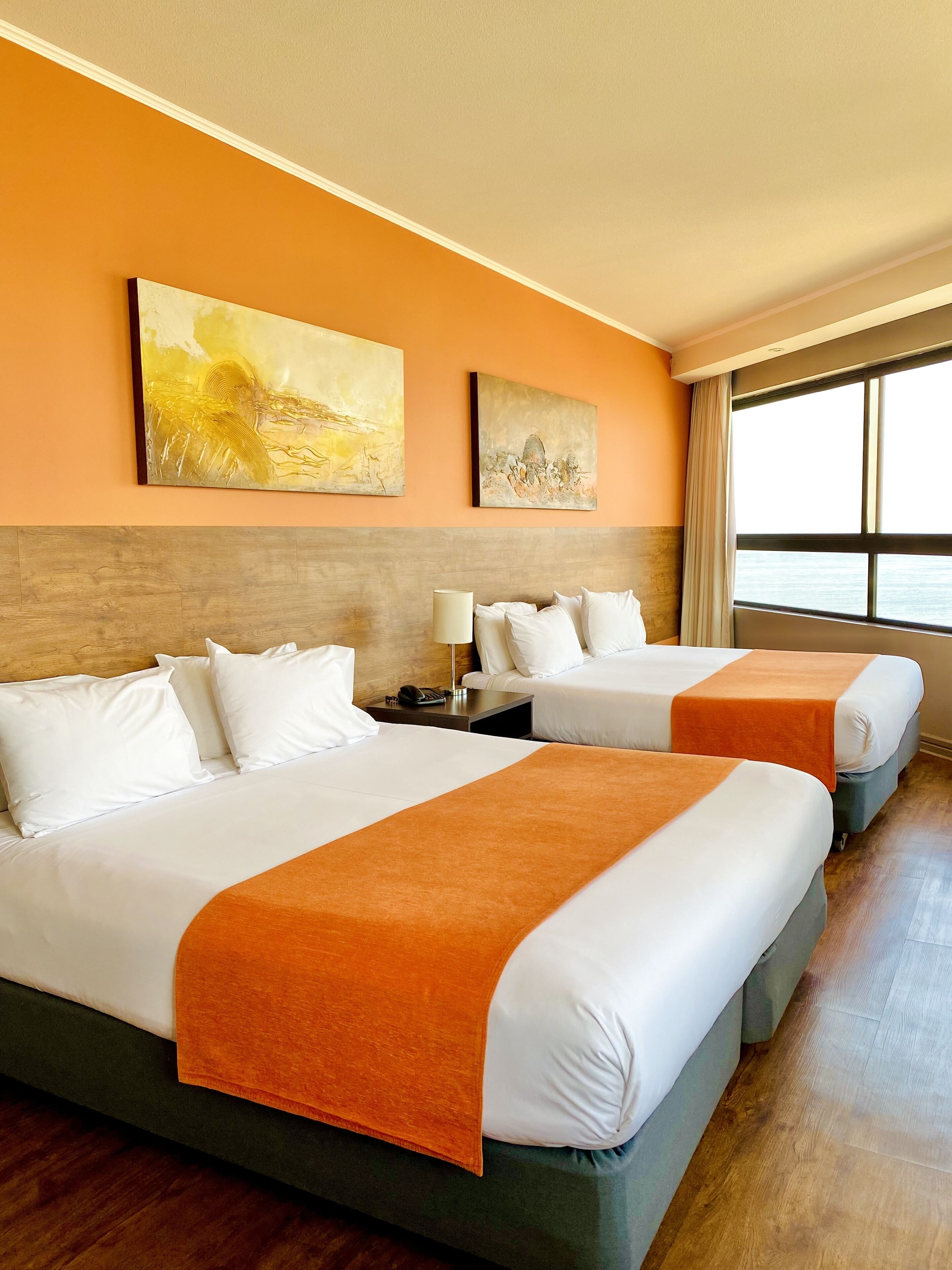A hotel room with two beds, a wooden headboard, a nightstand with a lamp, and artwork on the wall.