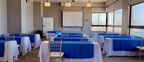 Un salón de conferencias con mesas dispuestas para una reunión, una pizarra y una pantalla de proyector.