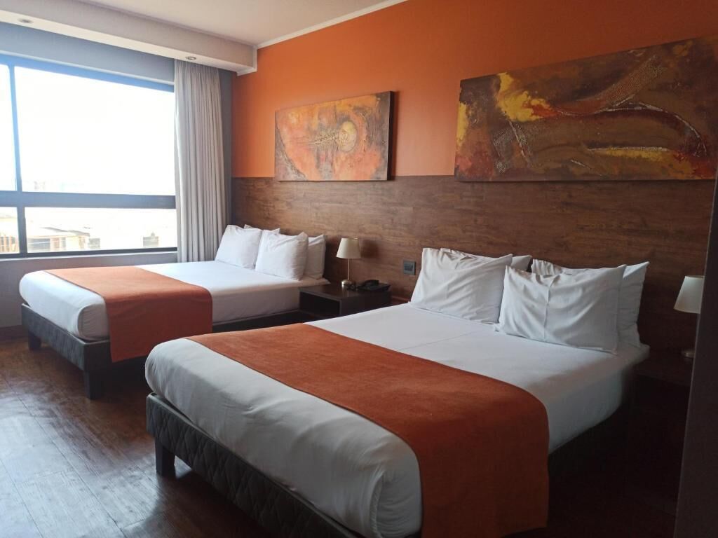 Habitación de hotel con dos camas, cada una con ropa de cama blanca y almohadones naranja, cabeceros de madera y un amplio ventanal con cortinas.