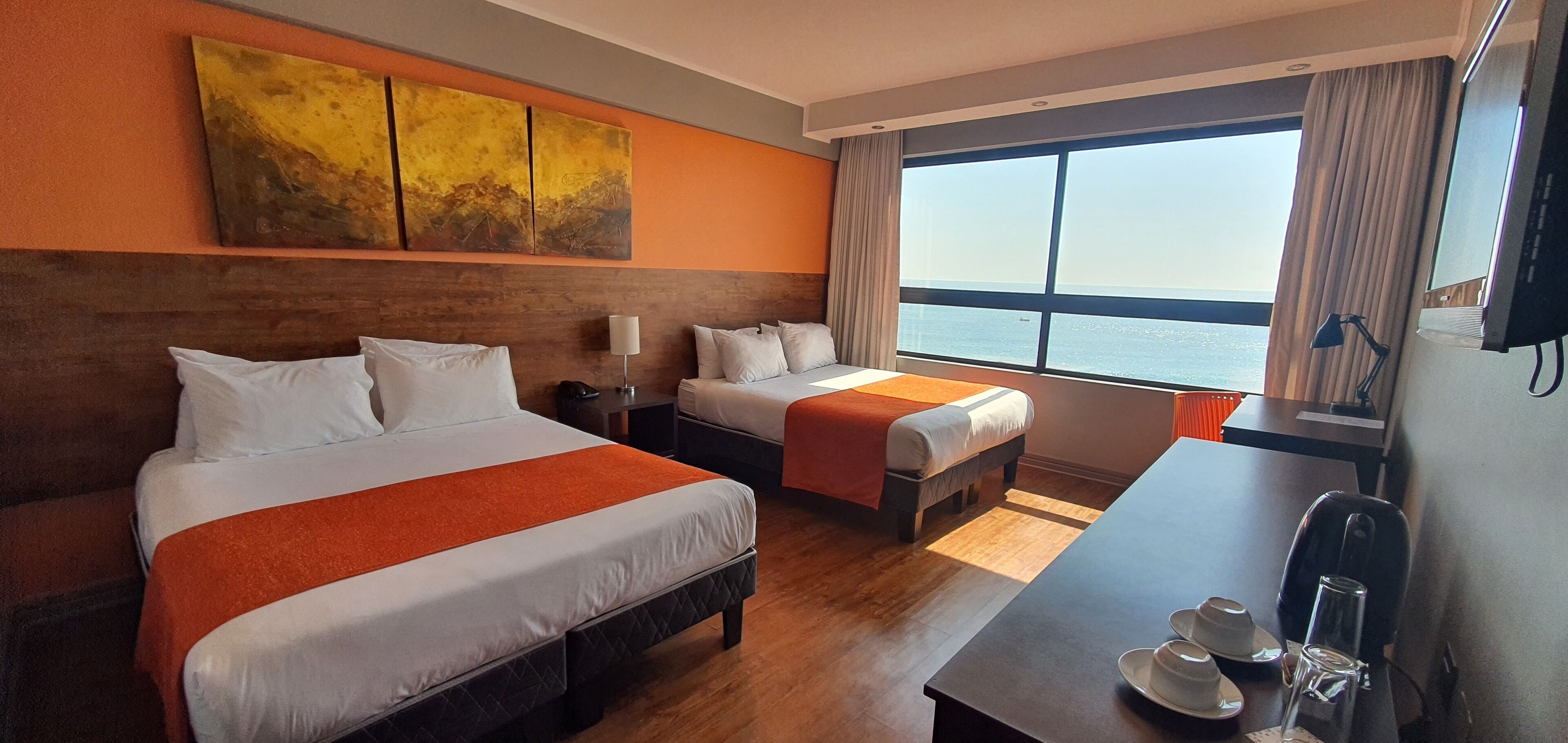 A hotel room with two beds, a large window with a view of the sea, a desk with a chair, and a TV on the wall.