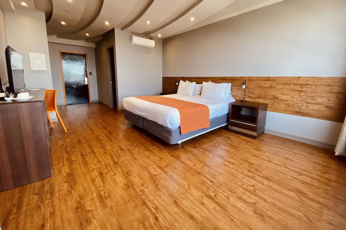 A modern hotel room with a wooden floor, a large bed with white linens, a wooden headboard, and a desk with a chair.
