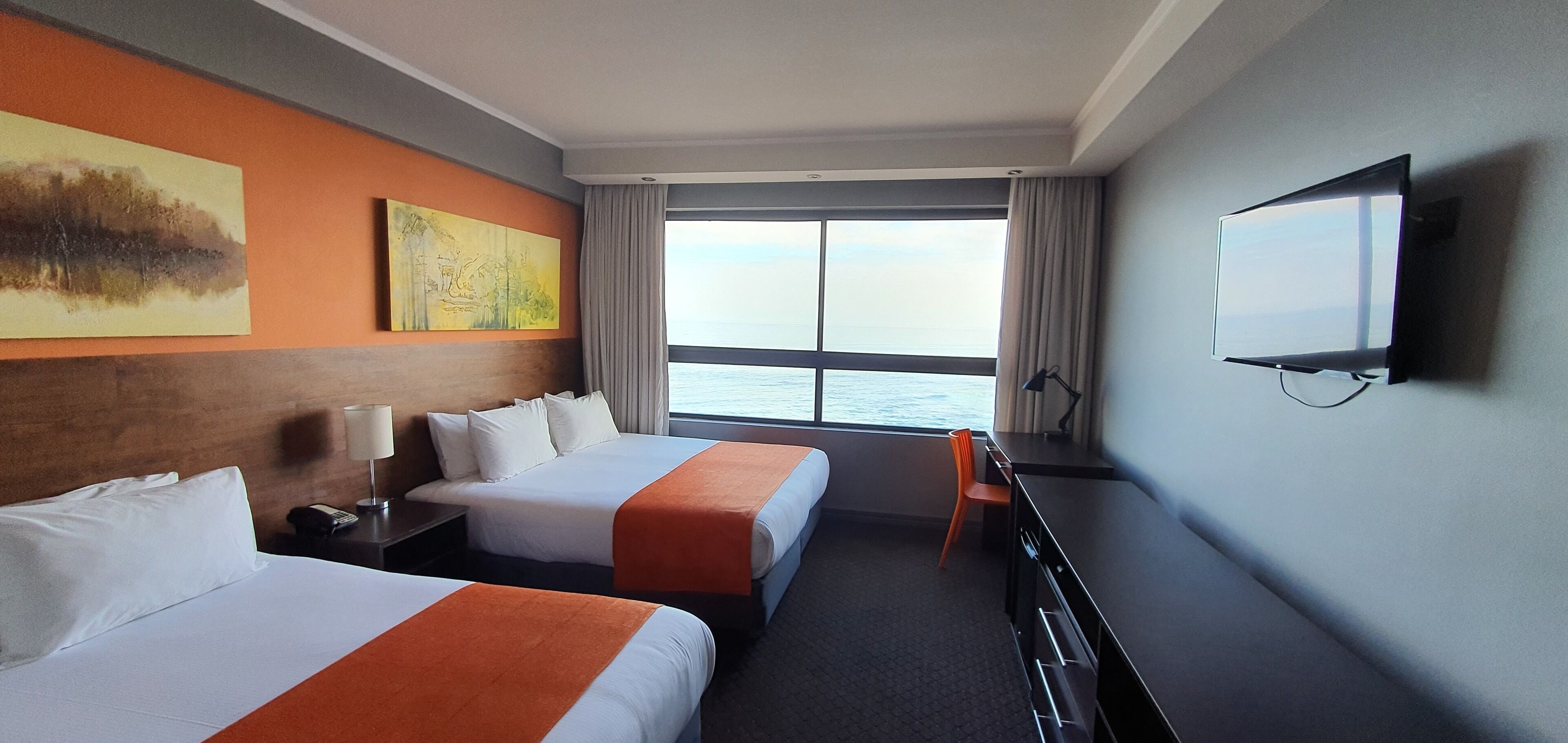 A hotel room with two beds, a desk, a chair, a TV, and a large window with a view of the sea.