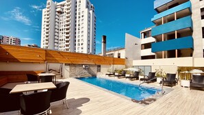 Un área de piscina en la azotea con un deck de madera, sillones de descanso y una mesa de comedor.