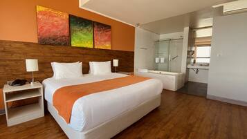 Una habitación de hotel moderna con una cama grande, una mesita de noche con lámpara, zona de ducha y baño con espejo.