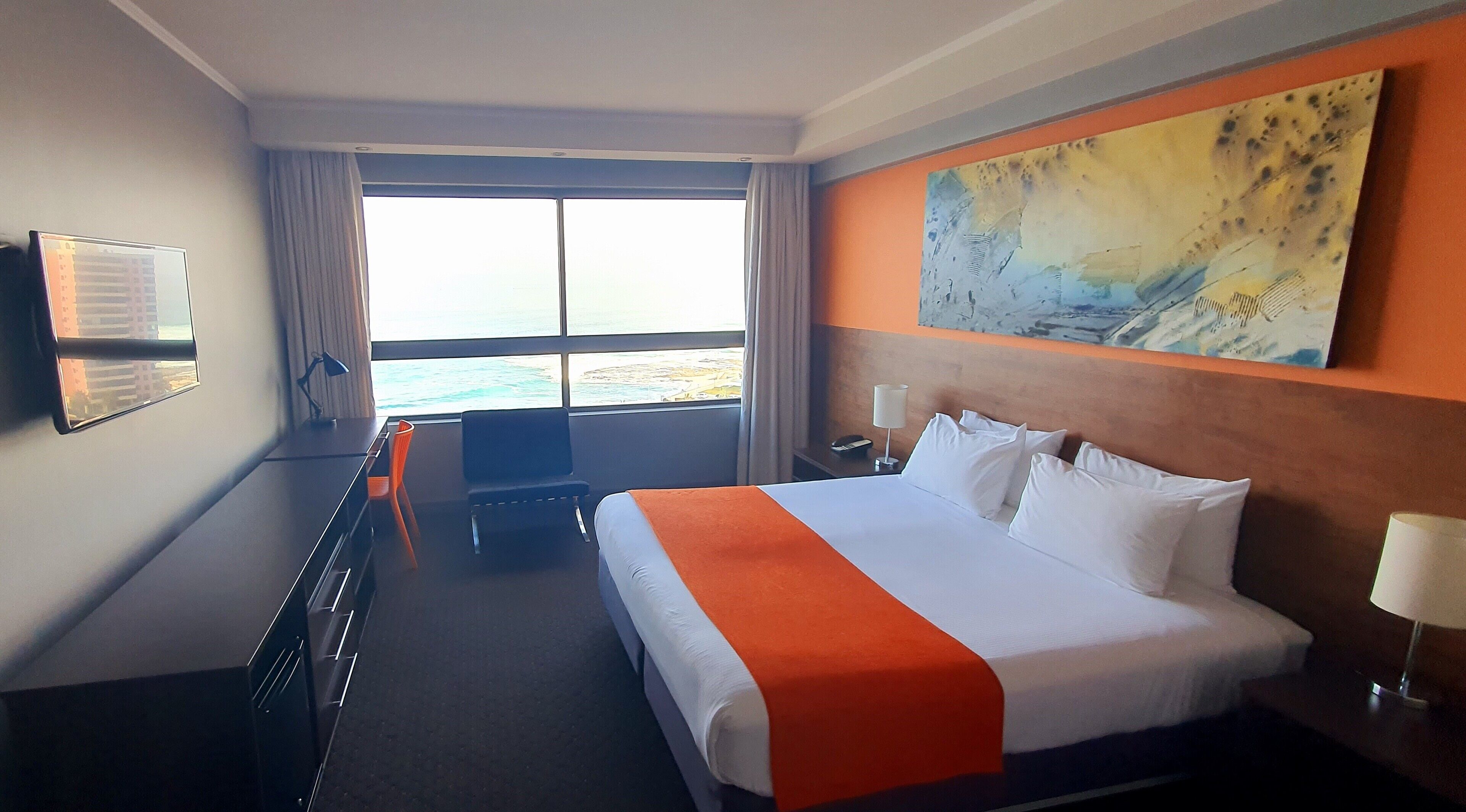 Quarto de hotel com cama, secretária, cadeira, televisão e grande janela com vista para o mar.