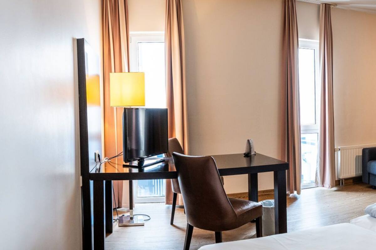 A hotel room with a bed, a desk with a chair, a sofa, a lamp, and a painting on the wall.