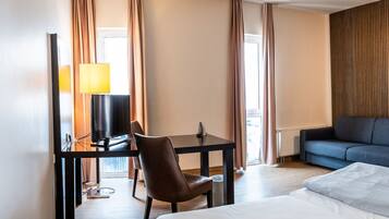 A hotel room with a bed, a desk with a chair, a sofa, a lamp, and a painting on the wall.