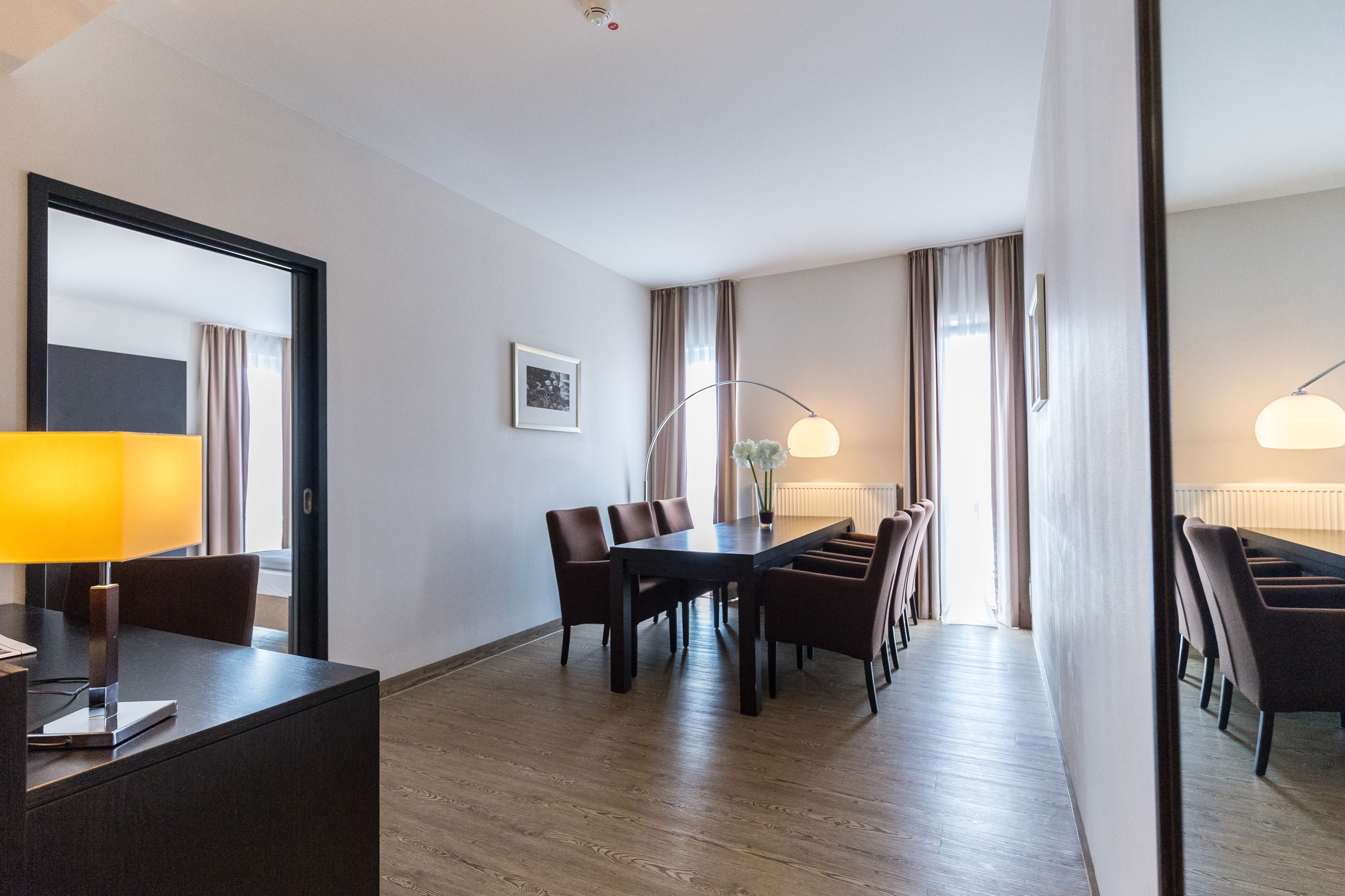 Une chambre d’hôtel équipée d’une table à manger, de chaises, d’un bureau avec une lampe et d’un grand miroir.