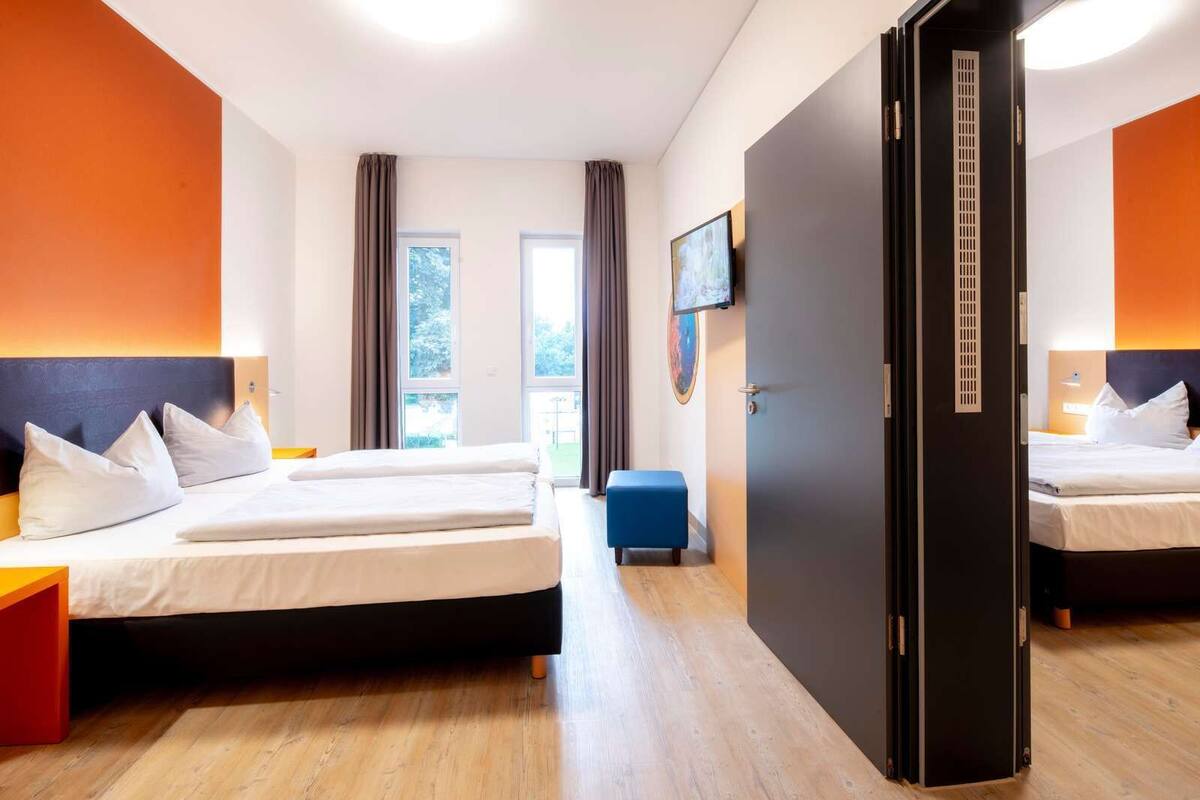 A hotel room with two beds, a blue ottoman, a large window, and a door with a vent.