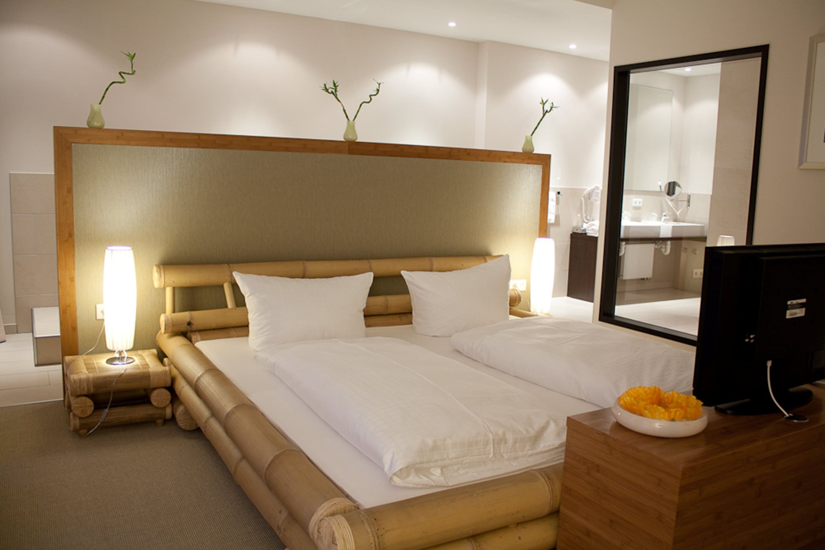 A hotel room with a large bed, a bedside table with a lamp, a wooden headboard, a mirror, and a television on a wooden dresser.