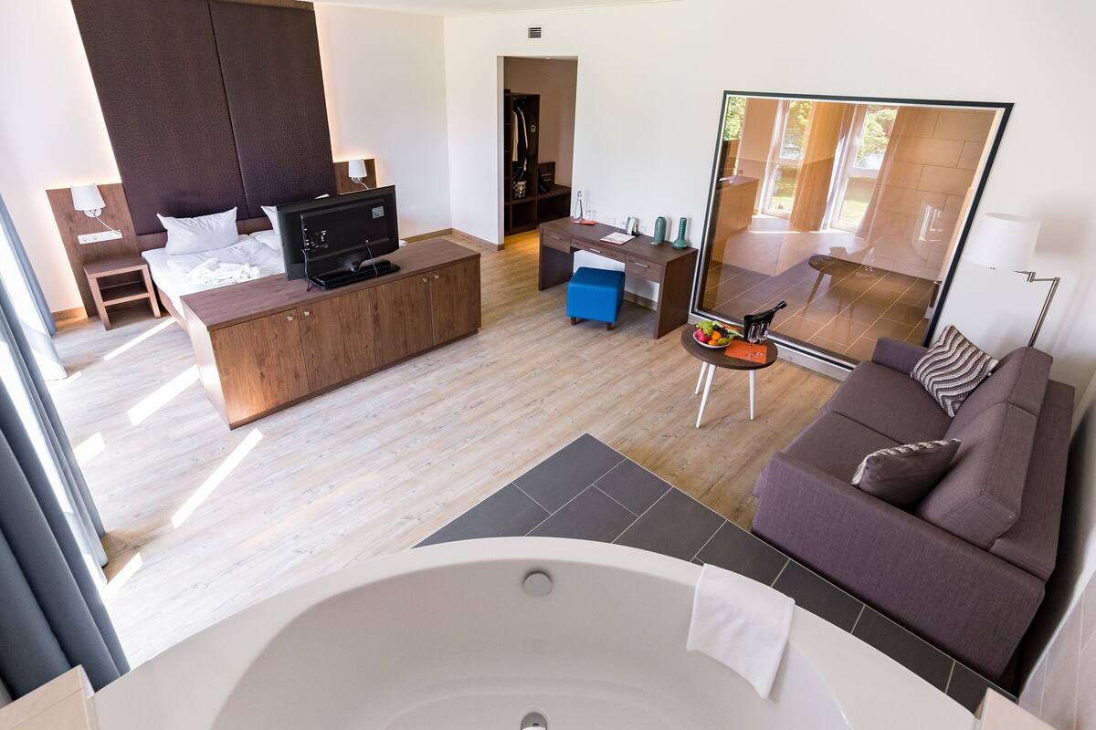 A modern hotel room with a large bed, a sofa, a desk, a TV, a mirror, and a bathtub.