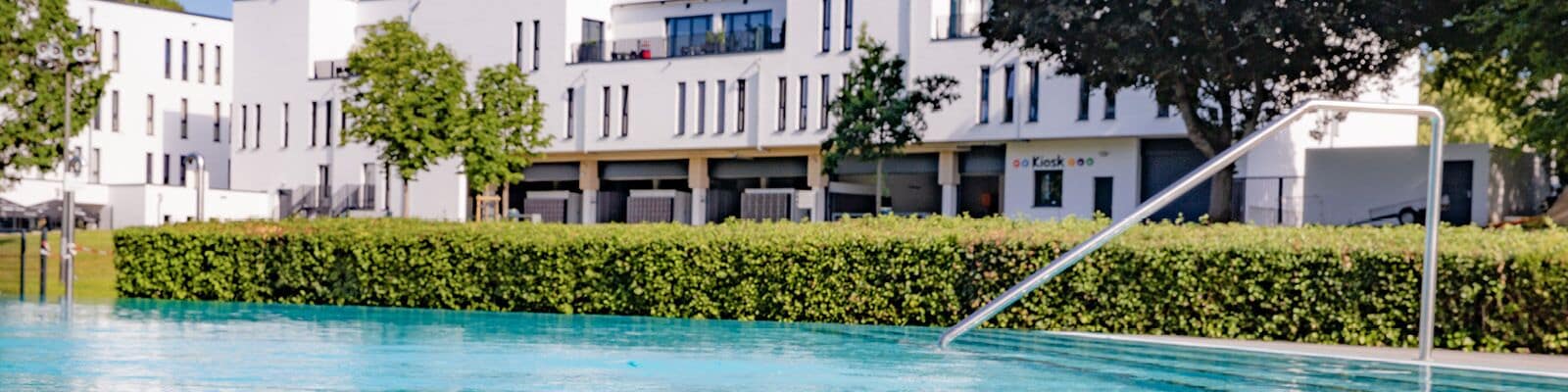 A swimming pool with a metal ladder, surrounded by a hedge and modern buildings in the background.