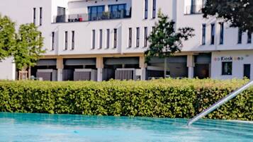 A swimming pool with a metal ladder, surrounded by a hedge and modern buildings in the background.