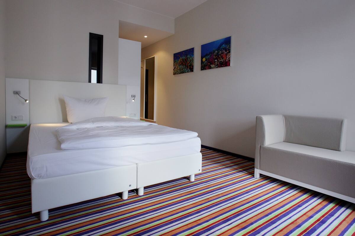 A modern hotel room with a large bed, a white sofa, colorful striped carpet, and two paintings on the wall.
