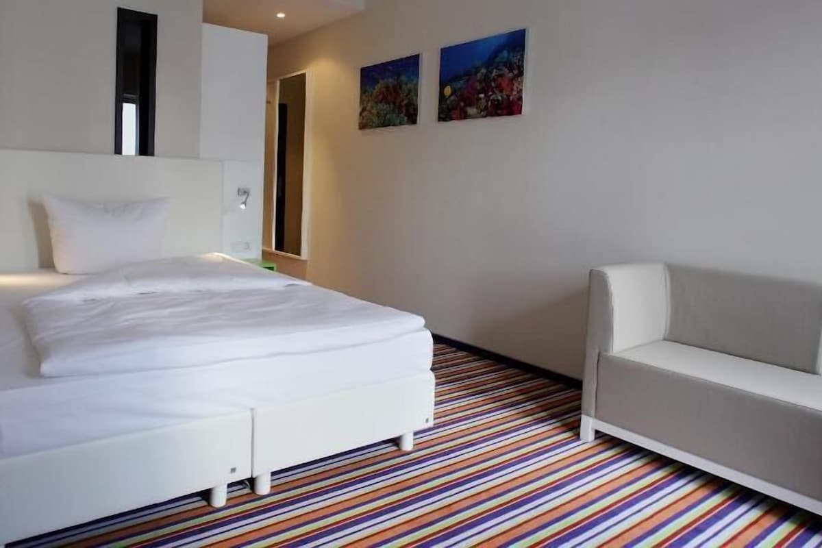 A hotel room with a bed, a white sofa, a striped carpet, and two wall-mounted paintings.