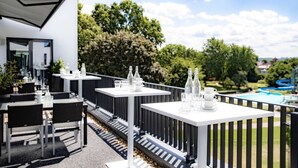 An outdoor dining area with white tables and chairs, surrounded by a black railing, overlooking a pool and greenery. - Friendly City Hotel Oktopus (Siegburg)