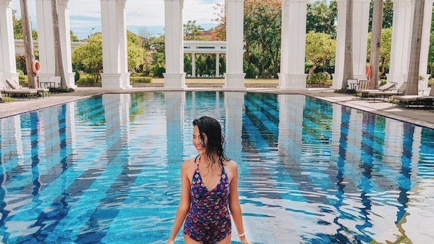 Una mujer con traje de baño estampado está de pie al borde de una piscina con agua azul cristalina, rodeada de columnas blancas y palmeras bajo un cielo luminoso.