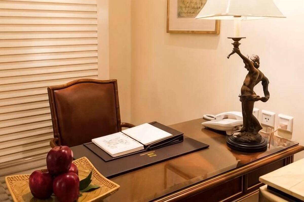 A hotel desk with a chair, a lamp, a telephone, a book, and a fruit basket.