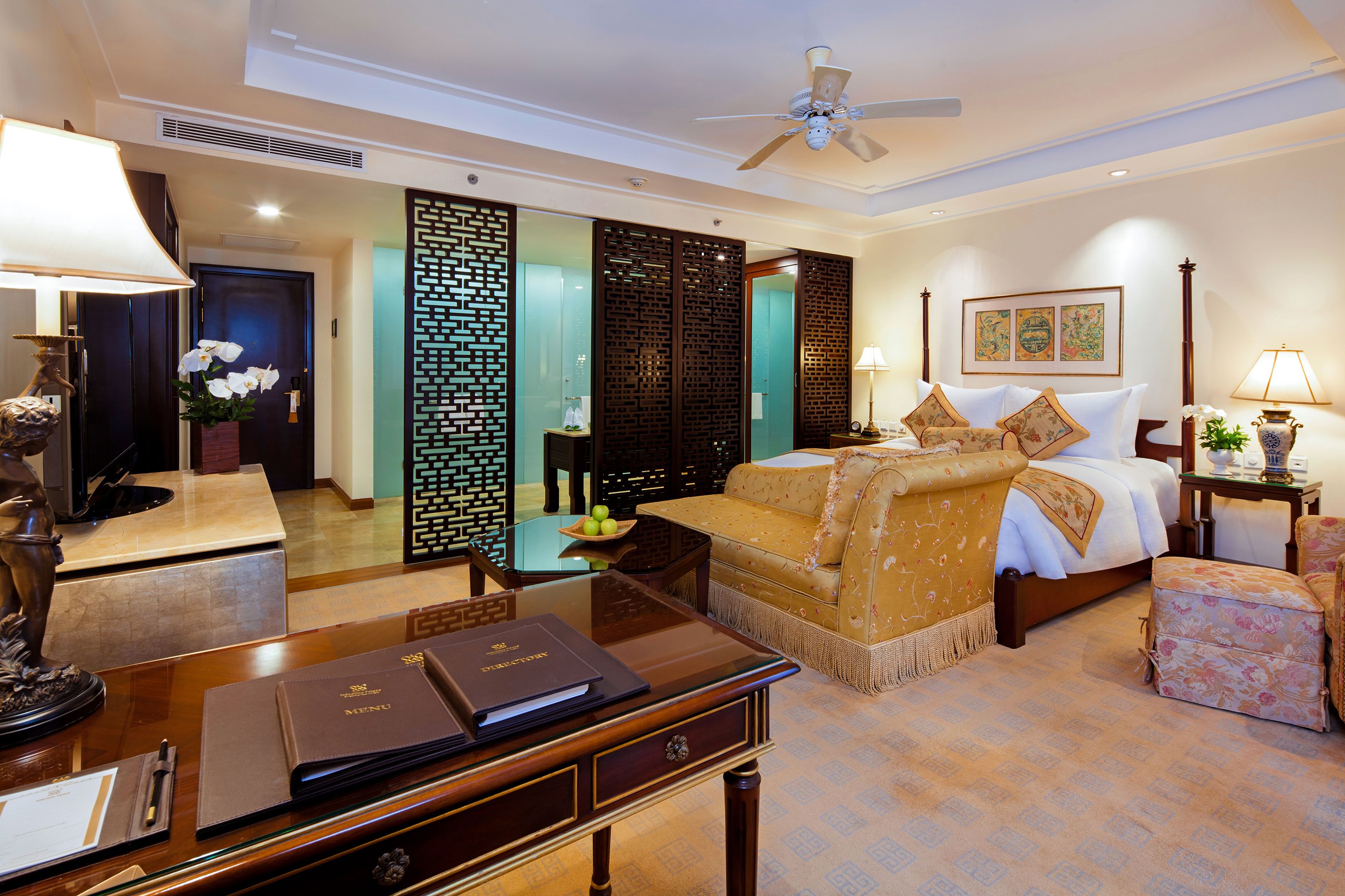 A hotel room with a bed, bedside table, lamp, decorative items, and a desk with a folder.