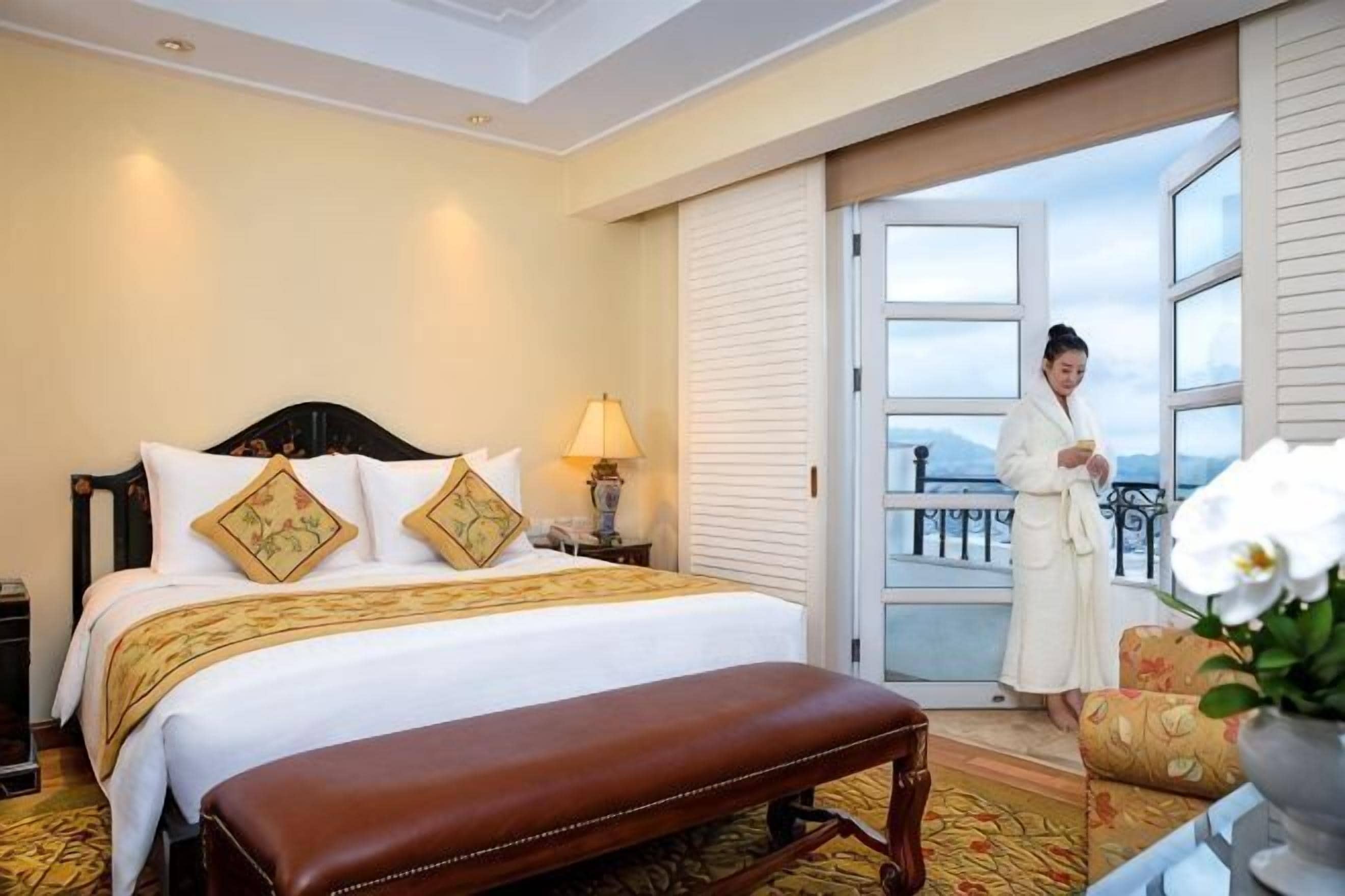A hotel room with a bed, a bench, a nightstand, a lamp, and a view of the outdoors.