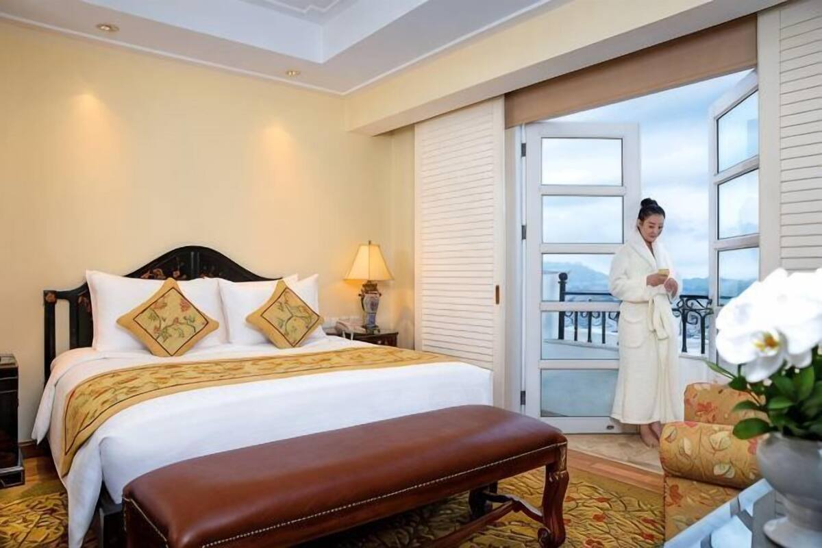 A hotel room with a bed, a bench, a nightstand, a lamp, and a view of the outdoors.