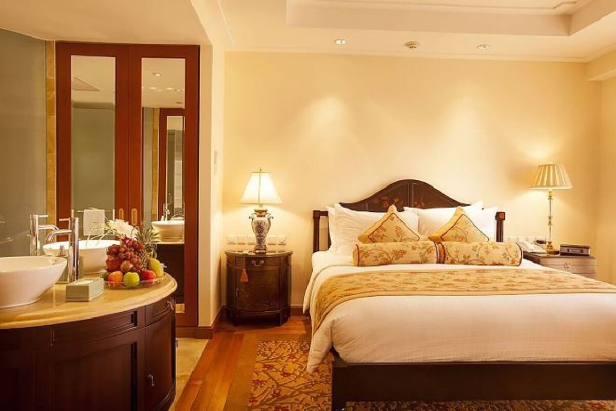 A hotel room with a large bed, a bedside table with a lamp, a vanity with a bowl sink, and a fruit bowl.