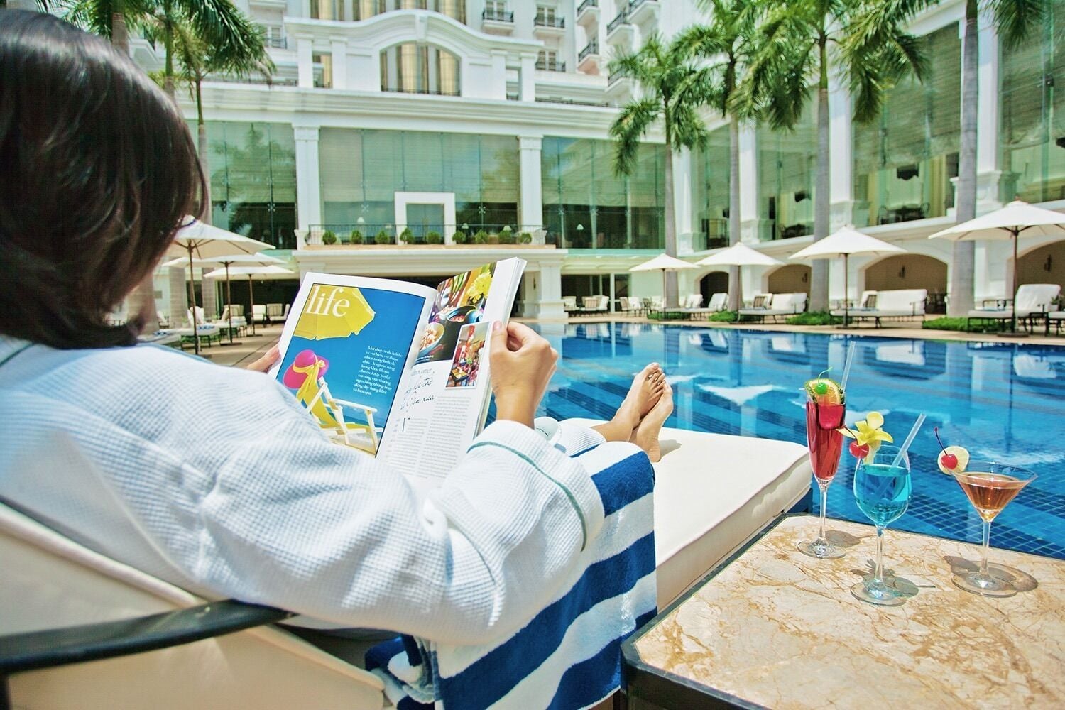 A person reading a magazine by a pool with lounge chairs and umbrellas in the background.