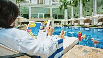 En person som sitter och läser en tidning vid en pool, med solstolar och parasoller i bakgrunden.