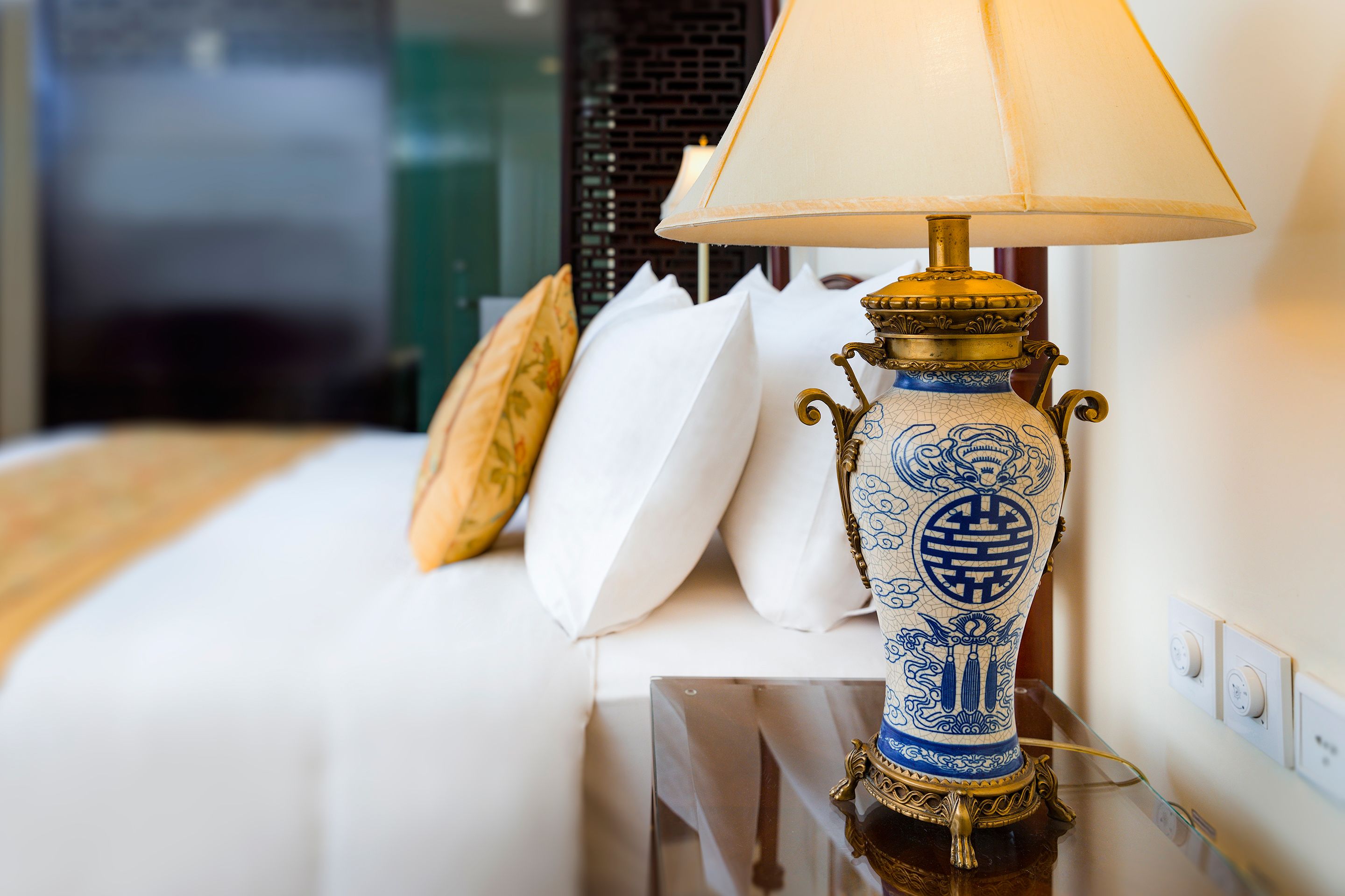 A bed with white bedding, a decorative vase lamp, and a wall with a power outlet.
