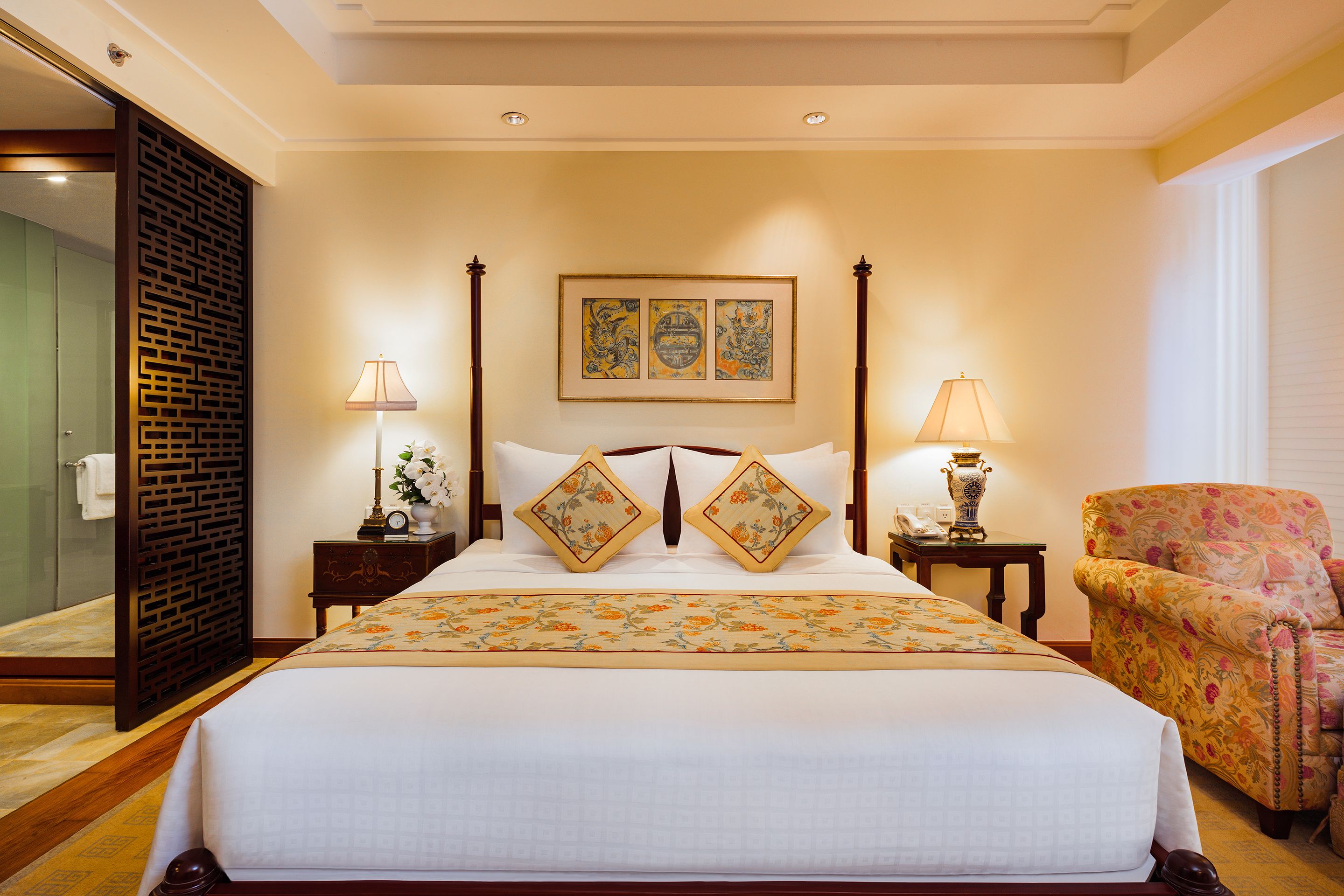 A large bed with a floral bedspread, two nightstands with lamps, a chair, and a wall with three framed artworks.