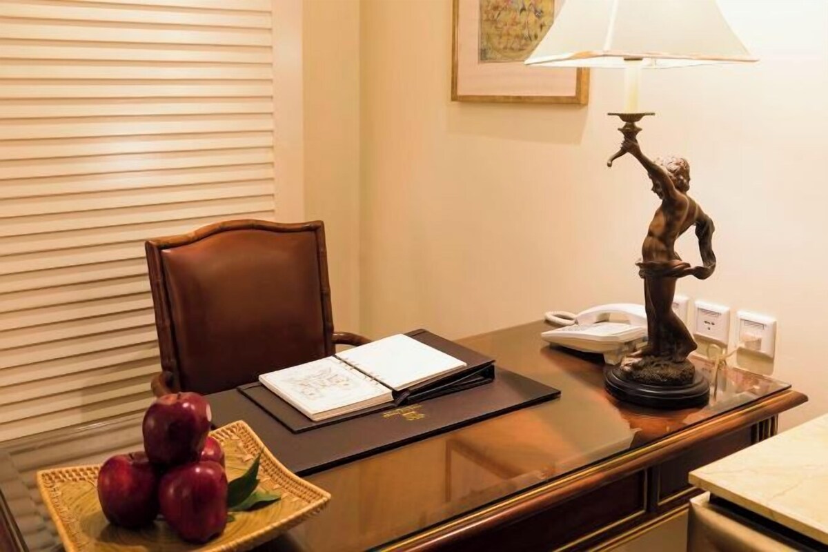 A hotel room with a wooden desk, a chair, a lamp, a telephone, a framed picture on the wall, and a basket with apples.