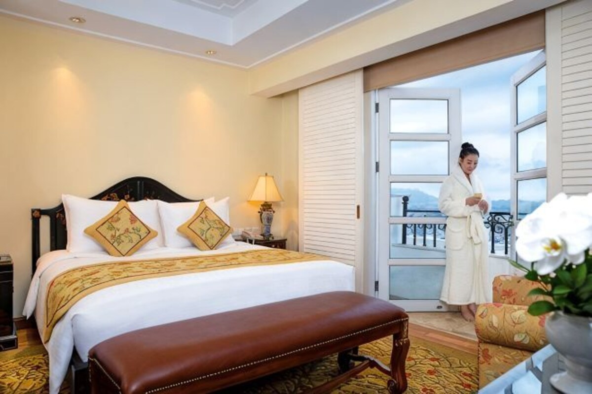 A hotel room with a bed, a bench, a nightstand, a lamp, and a view of the outdoors.