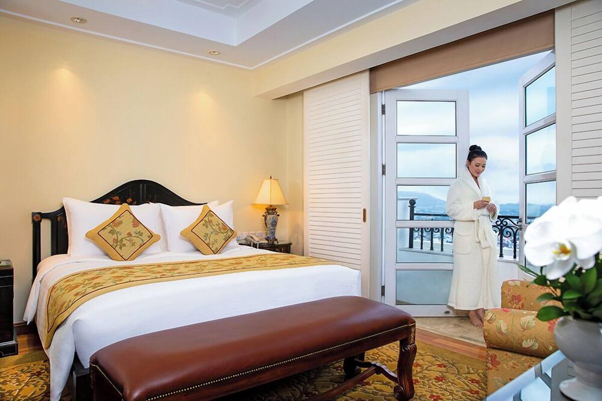 A hotel room with a large bed, a bench, a nightstand, a lamp, and a view of the outdoors.
