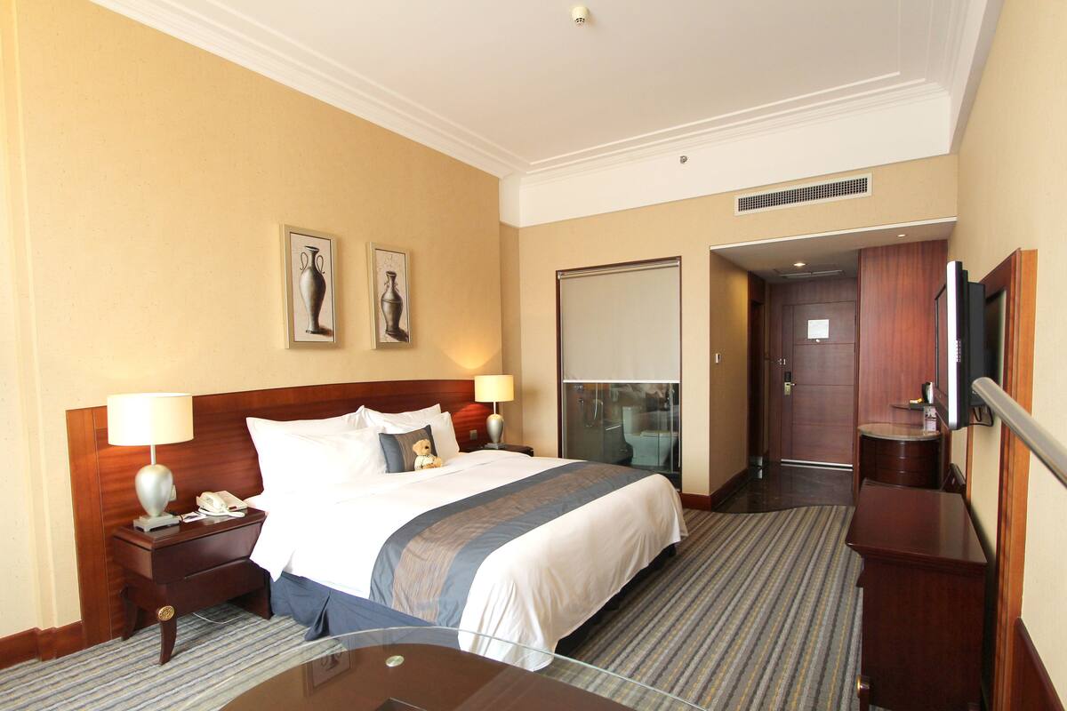 Une chambre d’hôtel avec un grand lit, une table de chevet avec une lampe, une cloison en verre, un téléviseur et une armoire en bois.