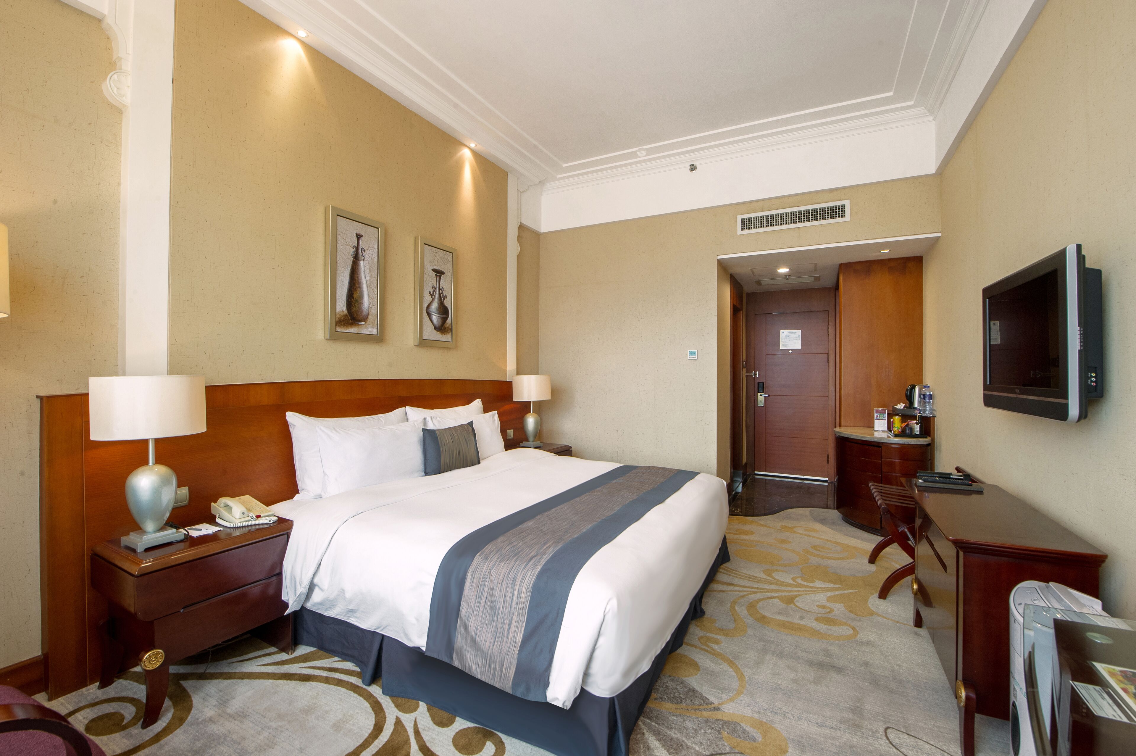 A hotel room with a large bed, bedside tables, a desk, a chair, a television, and a door leading to a bathroom.