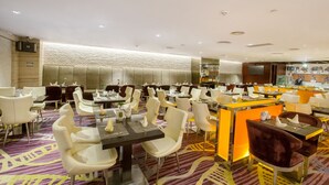 A modern restaurant with white and brown upholstered chairs, a patterned carpet, and a bar area with orange accents. - Asta Hotel (Shenzhen)