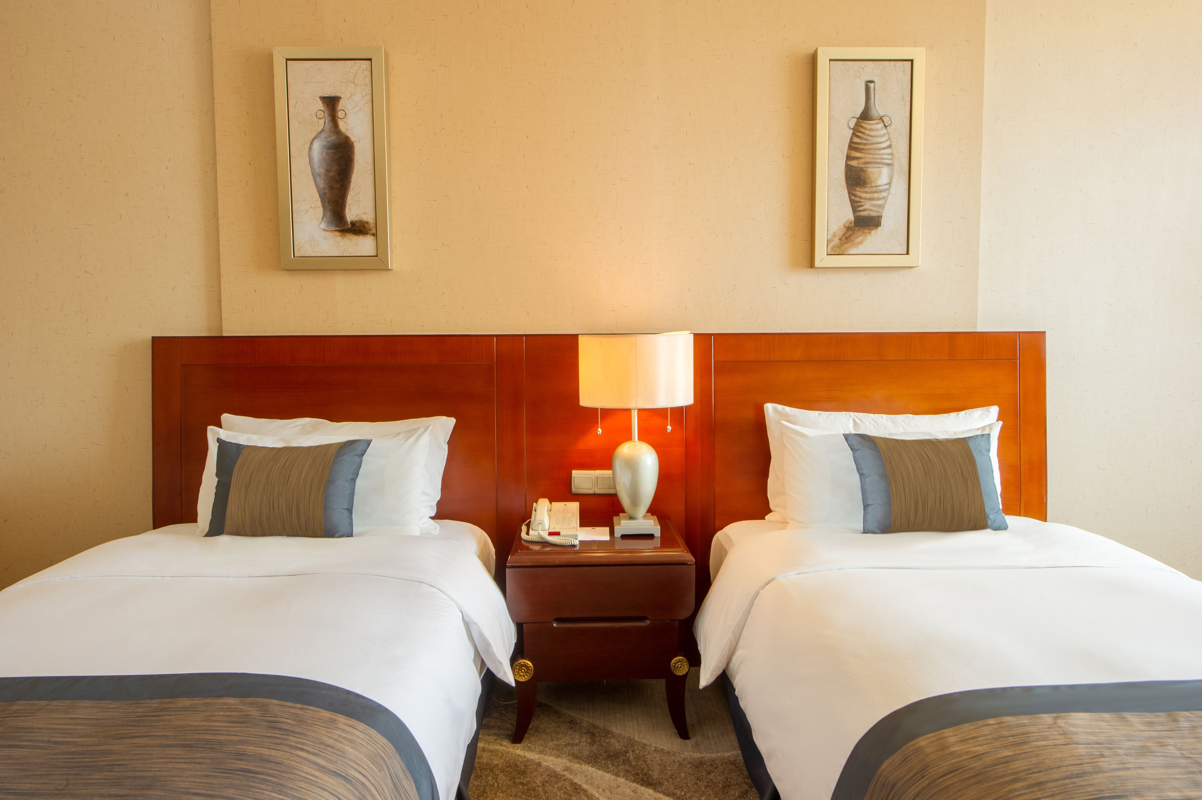 Une chambre d’hôtel avec deux lits, une tête de lit en bois, une table de chevet avec une lampe et deux œuvres d’art encadrées accrochées au mur.