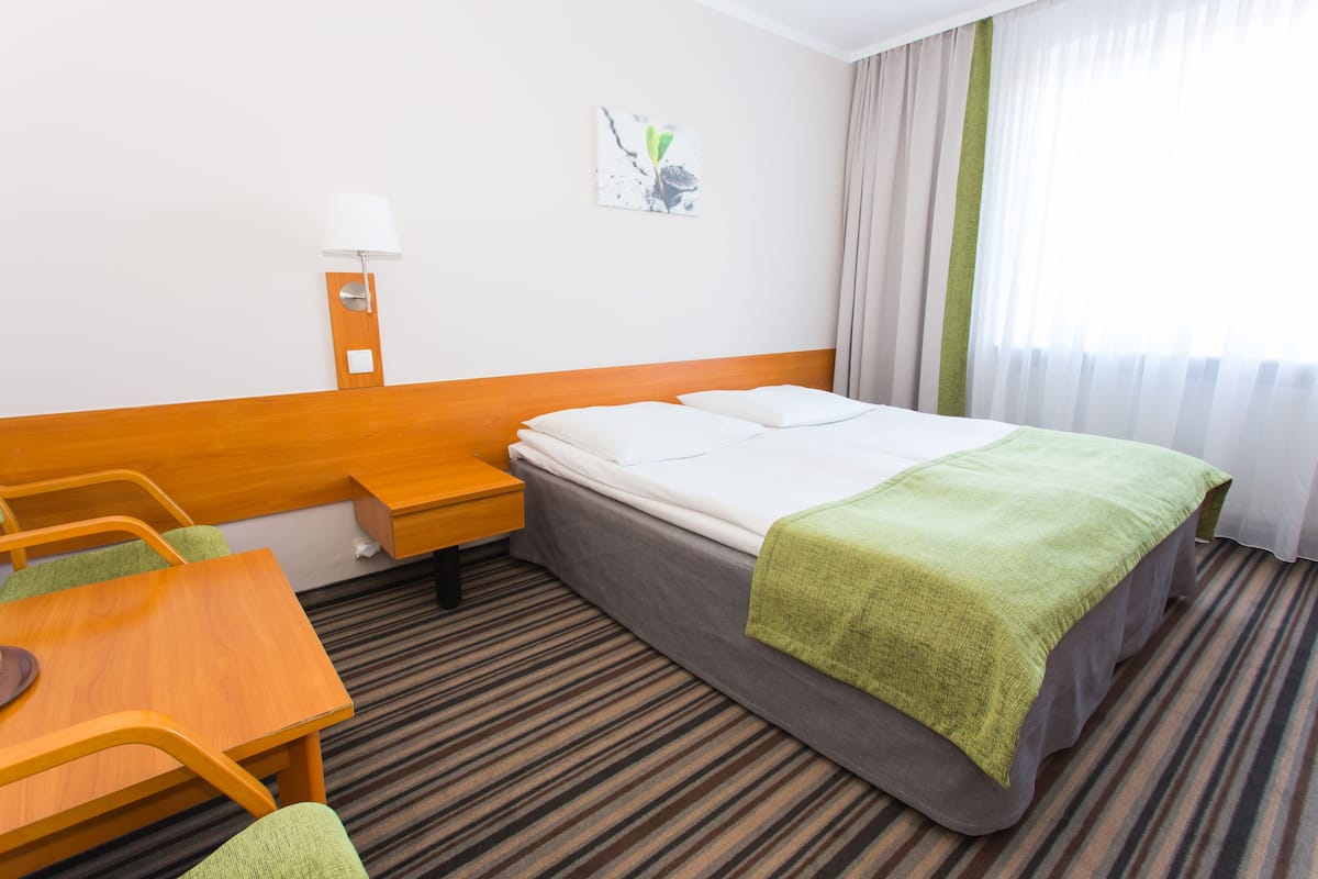 A hotel room with a bed, a desk, a chair, a lamp, and a window with curtains.