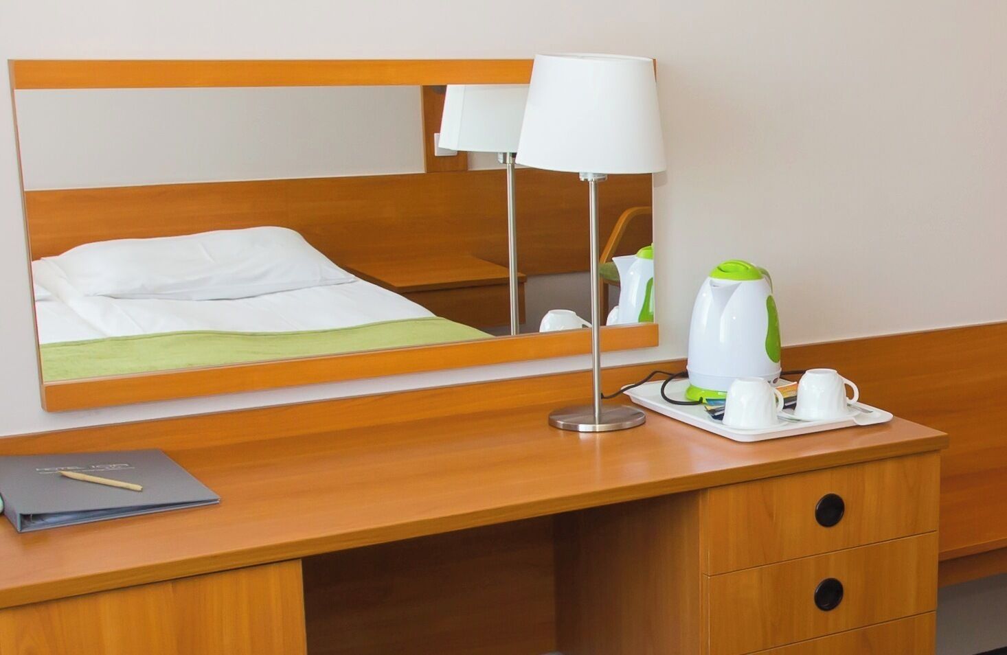 A hotel room with a wooden desk, a mirror, a lamp, a kettle, and a tray with cups and a book.